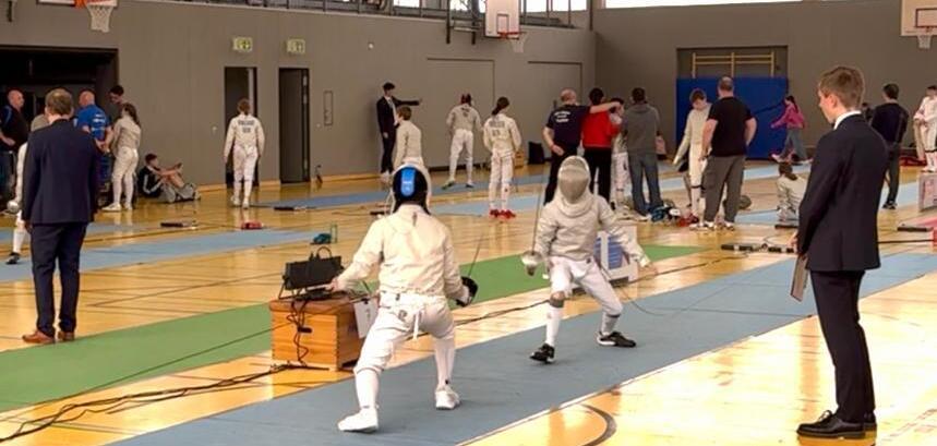 Fünf Fechter in Ausrüstung kämpfen auf einer bunten Fechtfläche in einer Sporthalle, Zuschauer beobachten das Geschehen.