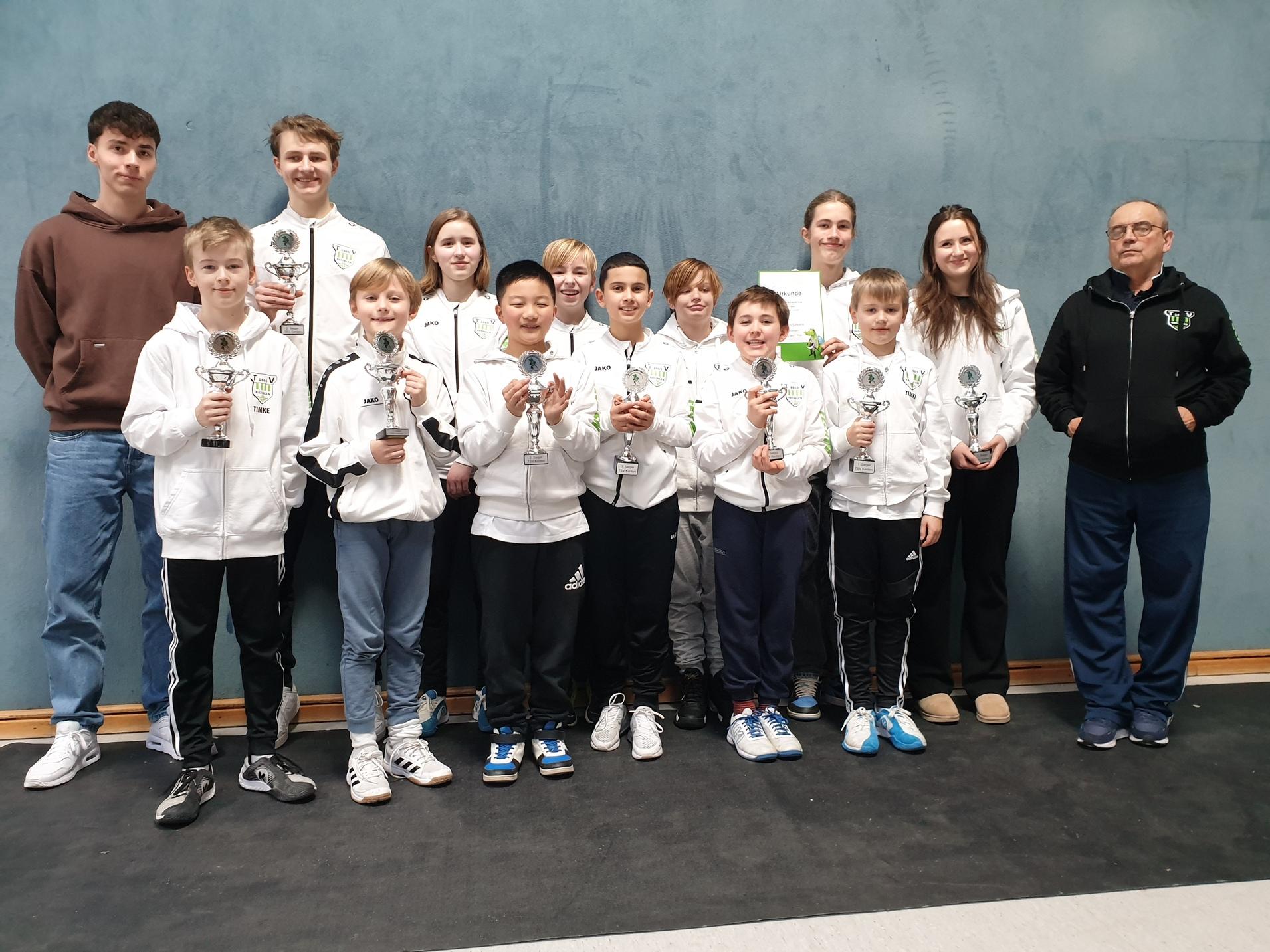 Gruppe von jungen Sportlern in weißen Jacken mit Pokalen, stehend vor einer hellblauen Wand.