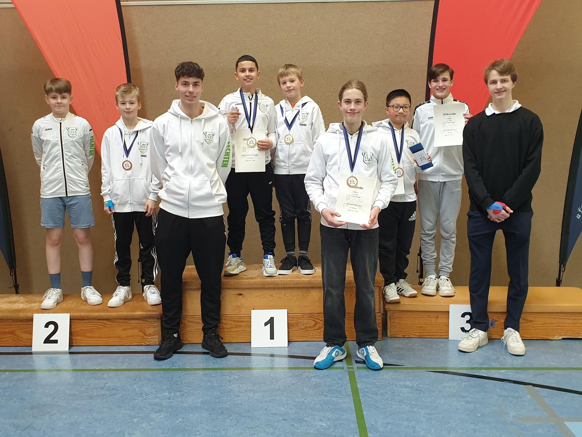 Gruppe von sechs Sportlern in Trainingsjacken auf dem Siegerpodest, mit Medaillen und Urkunden in einer Sporthalle.