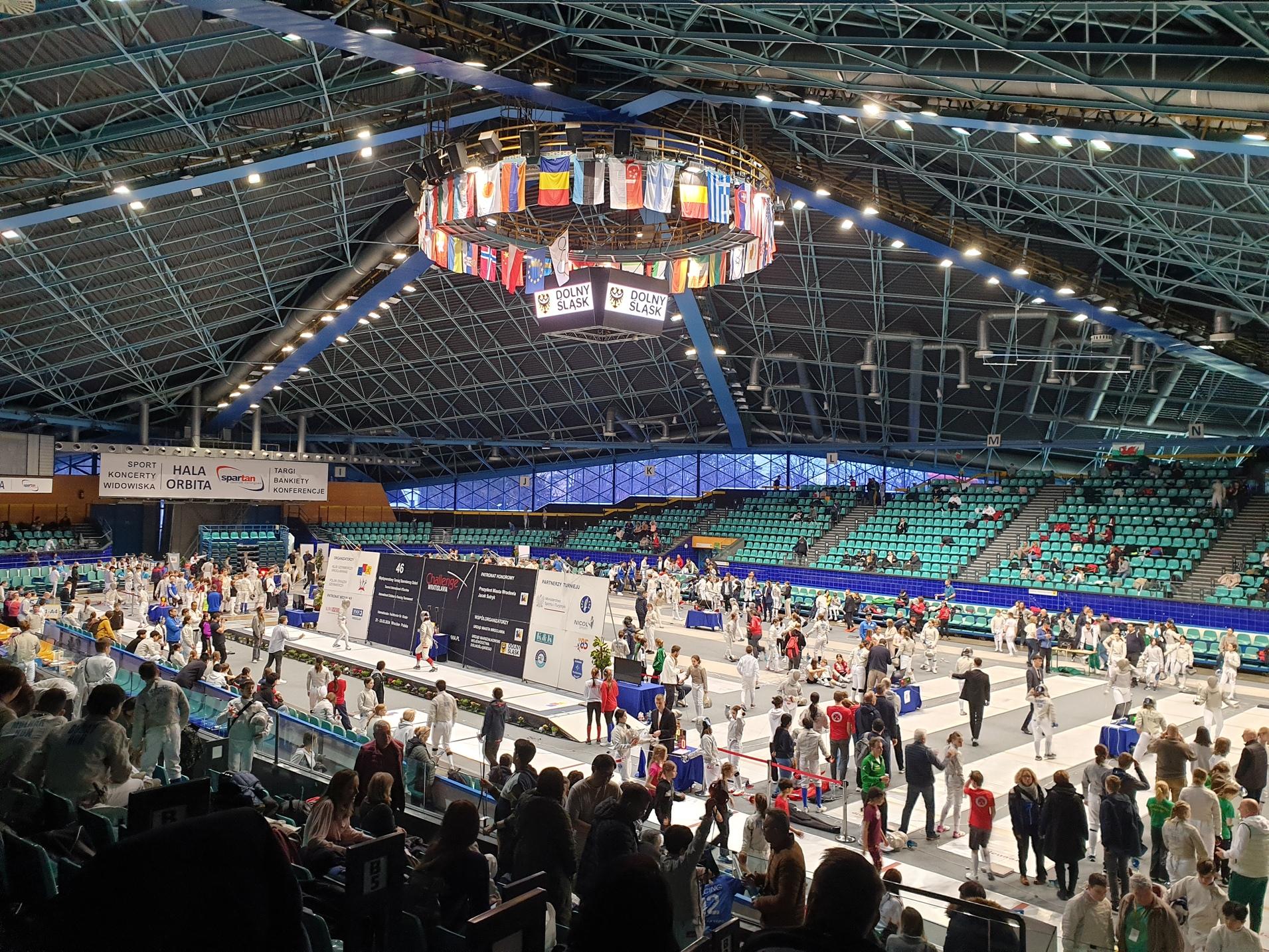 Innenansicht einer Sporthalle während eines Fechtturniers mit Teilnehmern, Zuschauern und Fahnen über dem Geschehen.