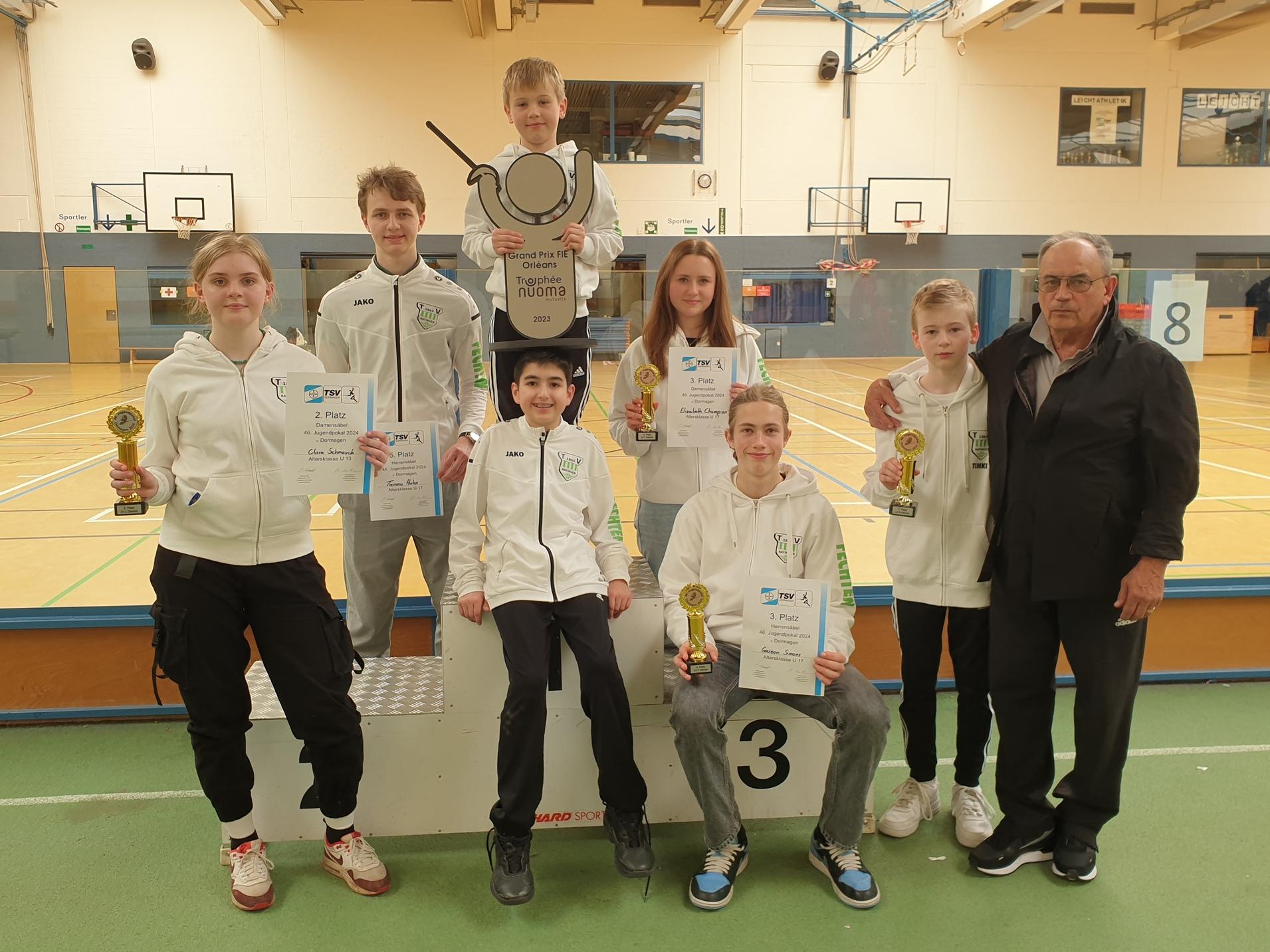 Gruppe von sechs Personen in Sportbekleidung mit Pokalen und Urkunden in einer Turnhalle nach einem Wettkampf.