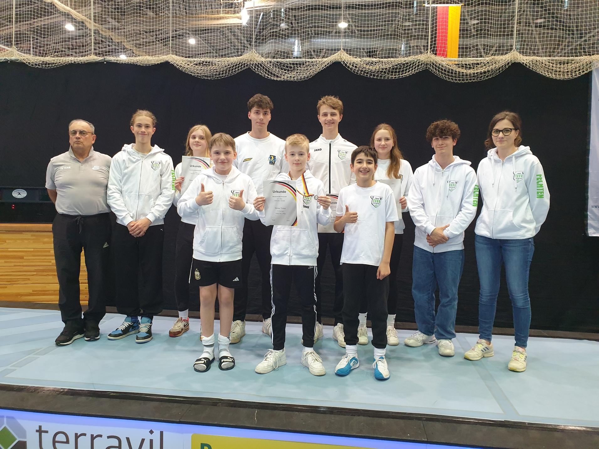 Gruppenbild von Sportlern in weißen Kapuzenjacken mit Urkunden und Medaillen auf einem Wettkampf-Podest.