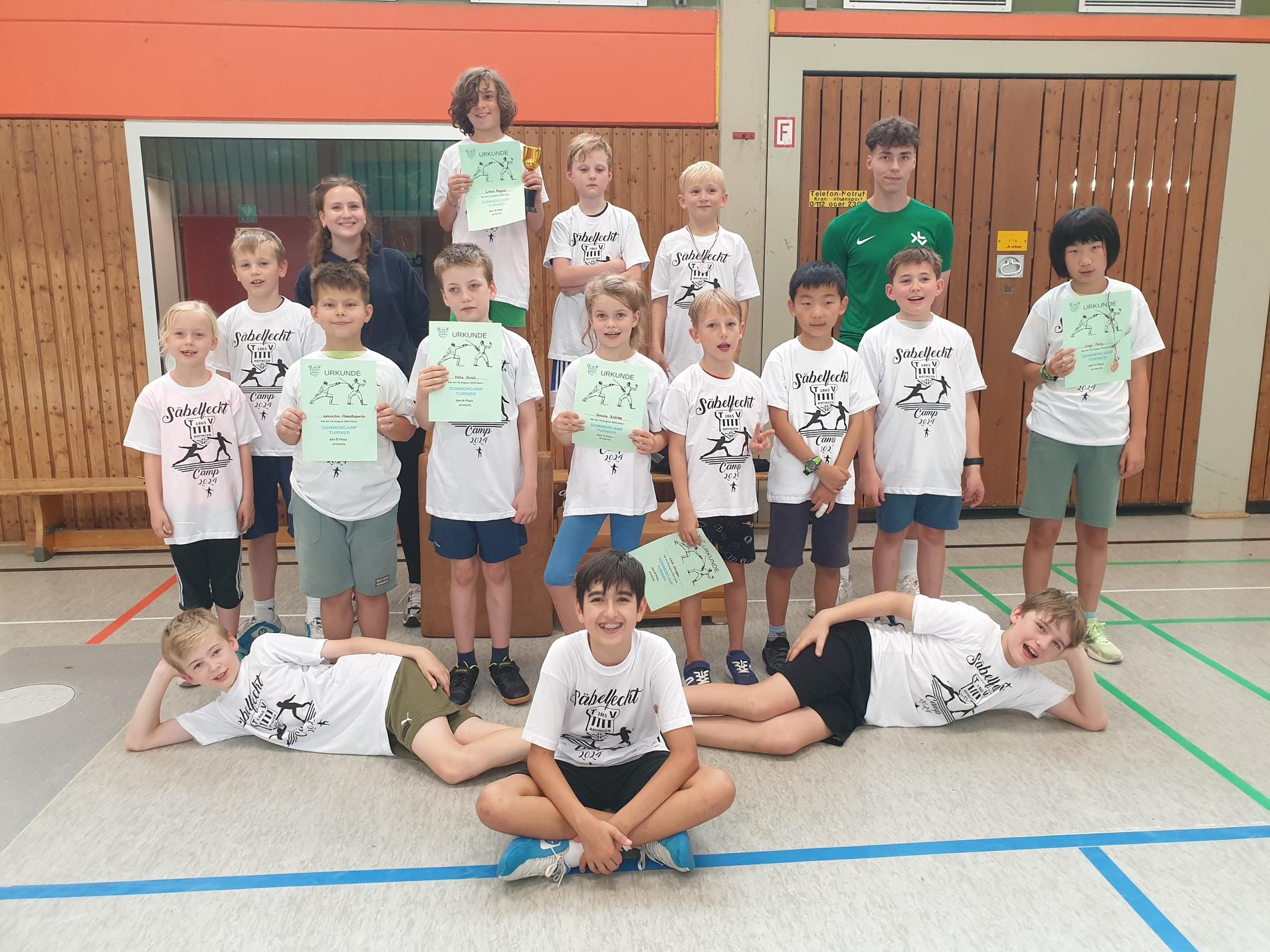 Gruppe von Kindern in T-Shirts mit Urkunden in einer Sporthalle, einige posieren, andere liegen entspannt auf dem Boden.