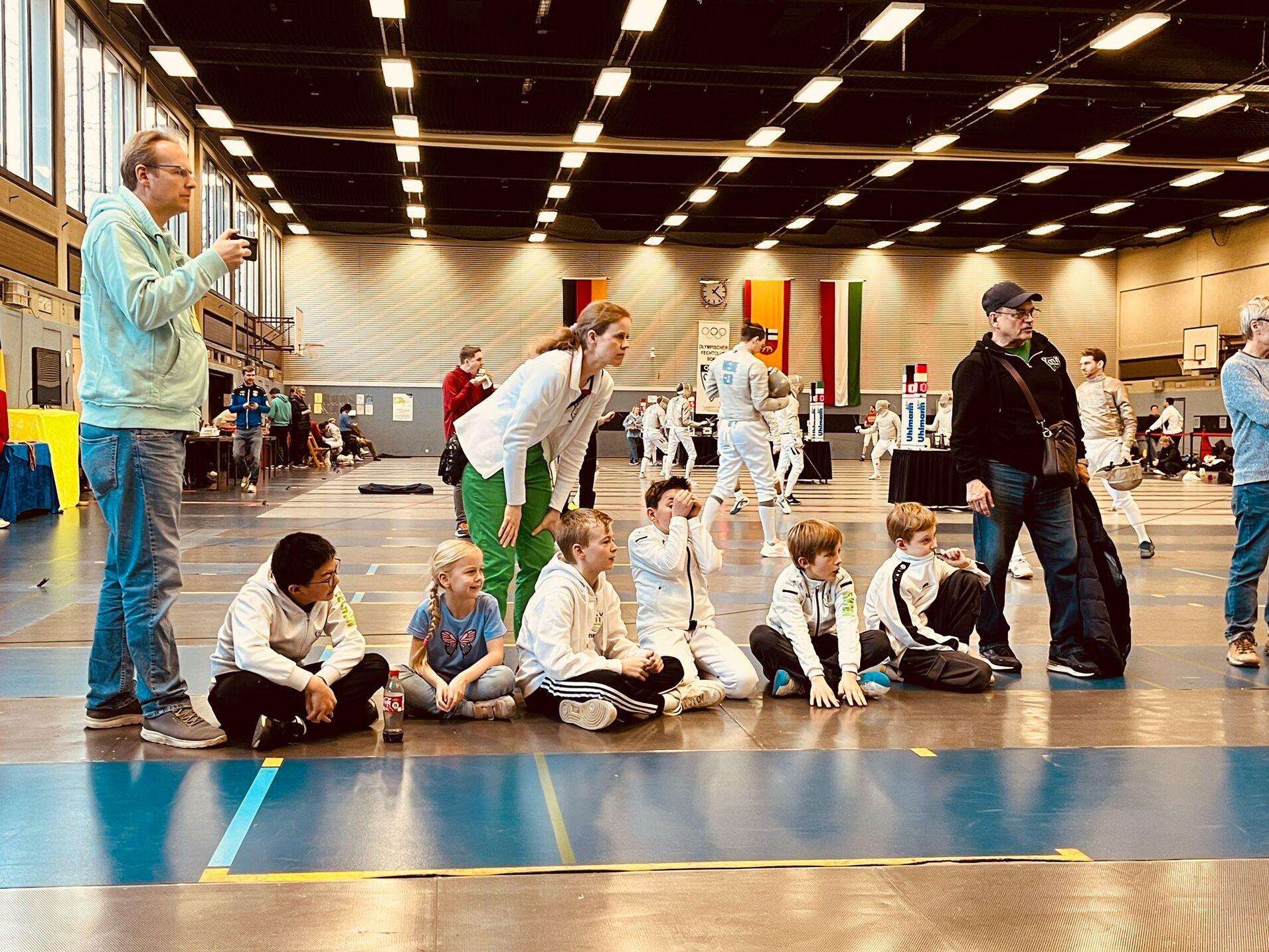 Szenen aus einem Fechtturnier: Zuschauer stehen und sitzen am Rand, während Kämpfer auf der Matte agieren.