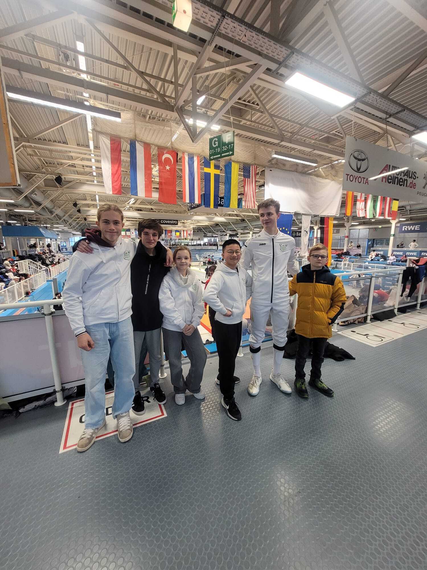 Gruppe von sechs Jugendlichen in Sportkleidung, stehend in einer Halle mit Flaggen im Hintergrund, Lächeln und Teamgeist.