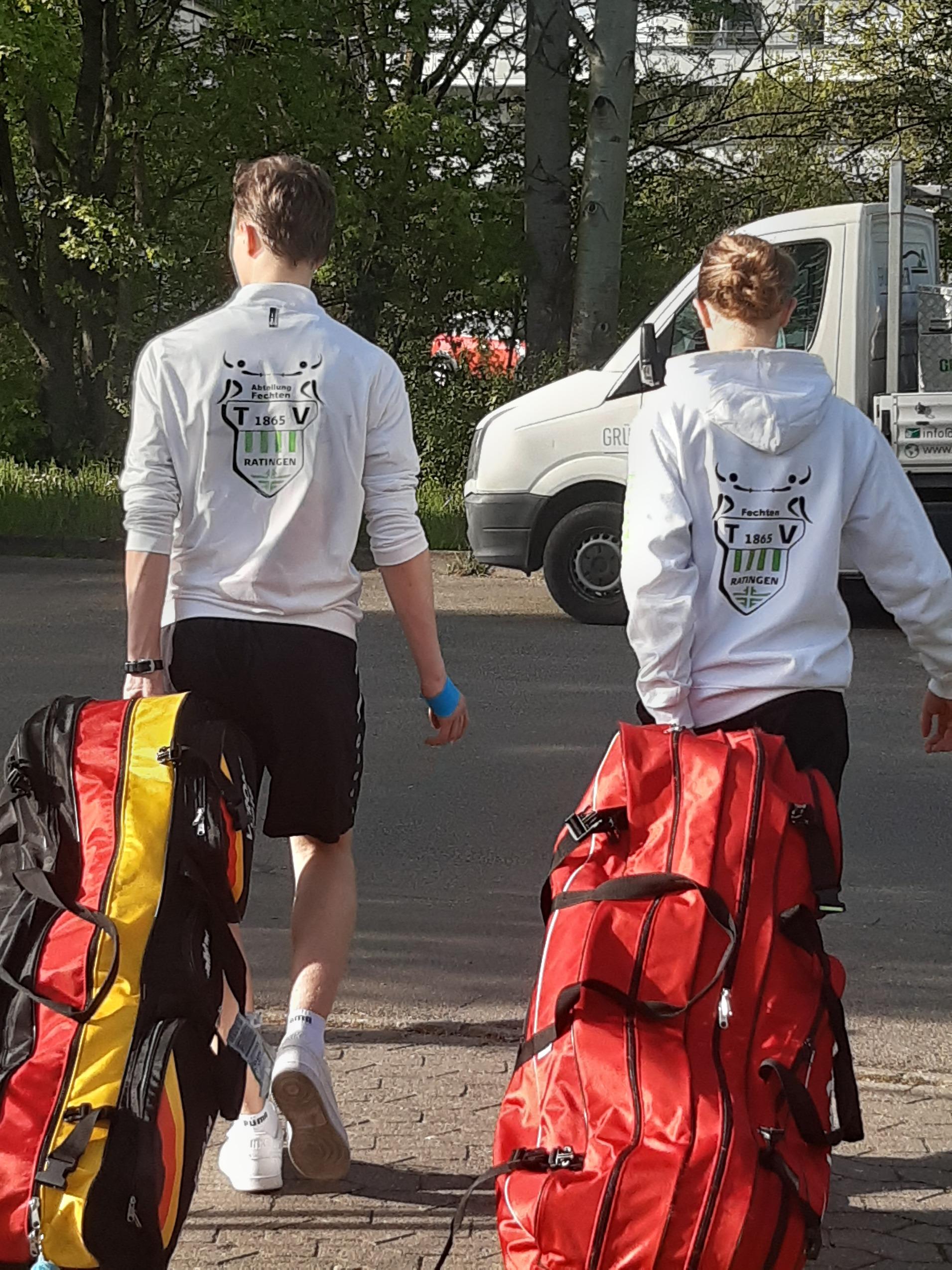 Zwei Sportler in weißen Kapuzenpullis ziehen große Sporttaschen auf einem Gehweg. Im Hintergrund stehen Bäume und ein Fahrzeug.