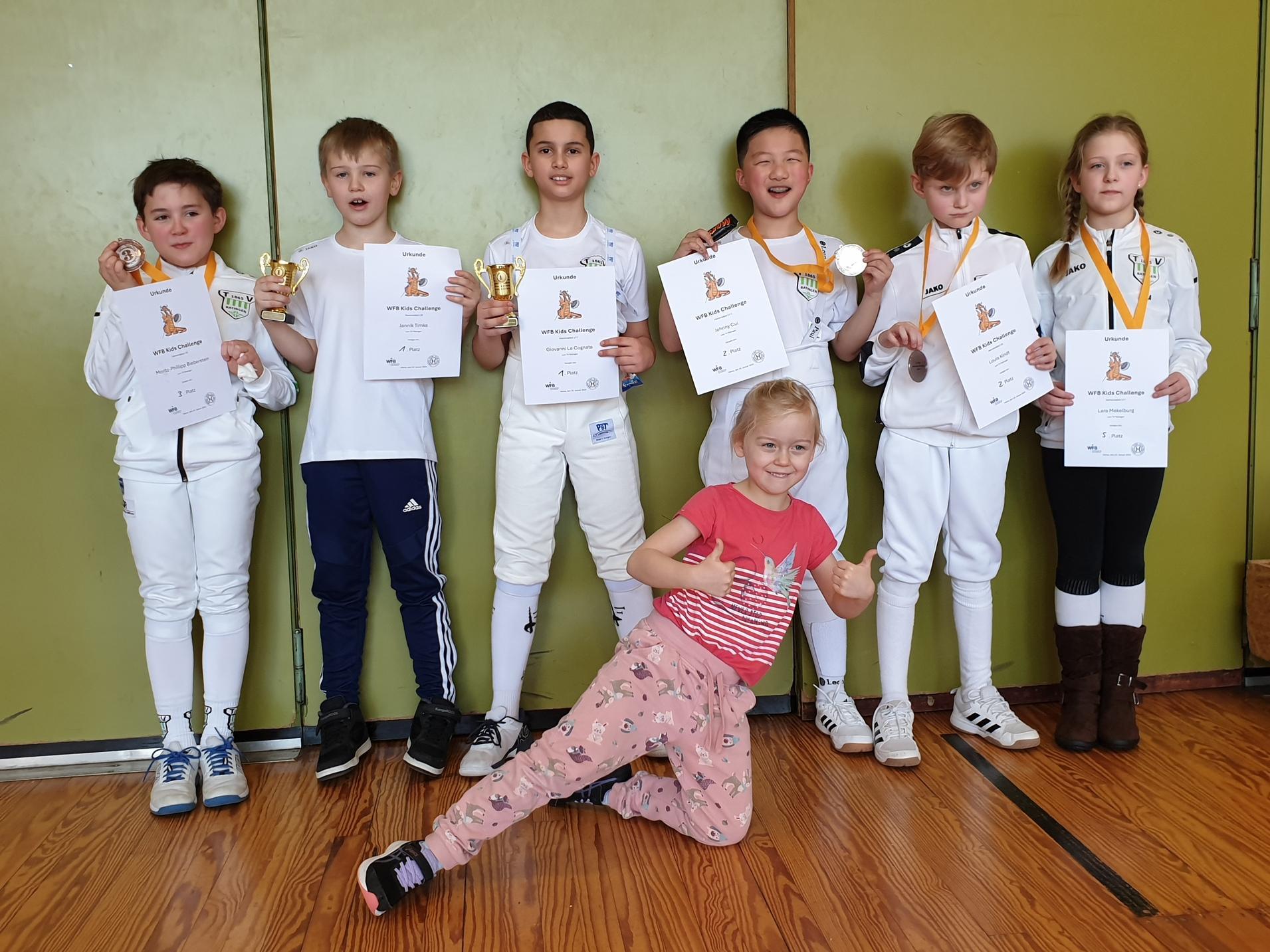 Sechs Kinder in Fechtkleidung halten Medaillen und Urkunden, während ein Mädchen im Vordergrund Daumen hoch zeigt.
