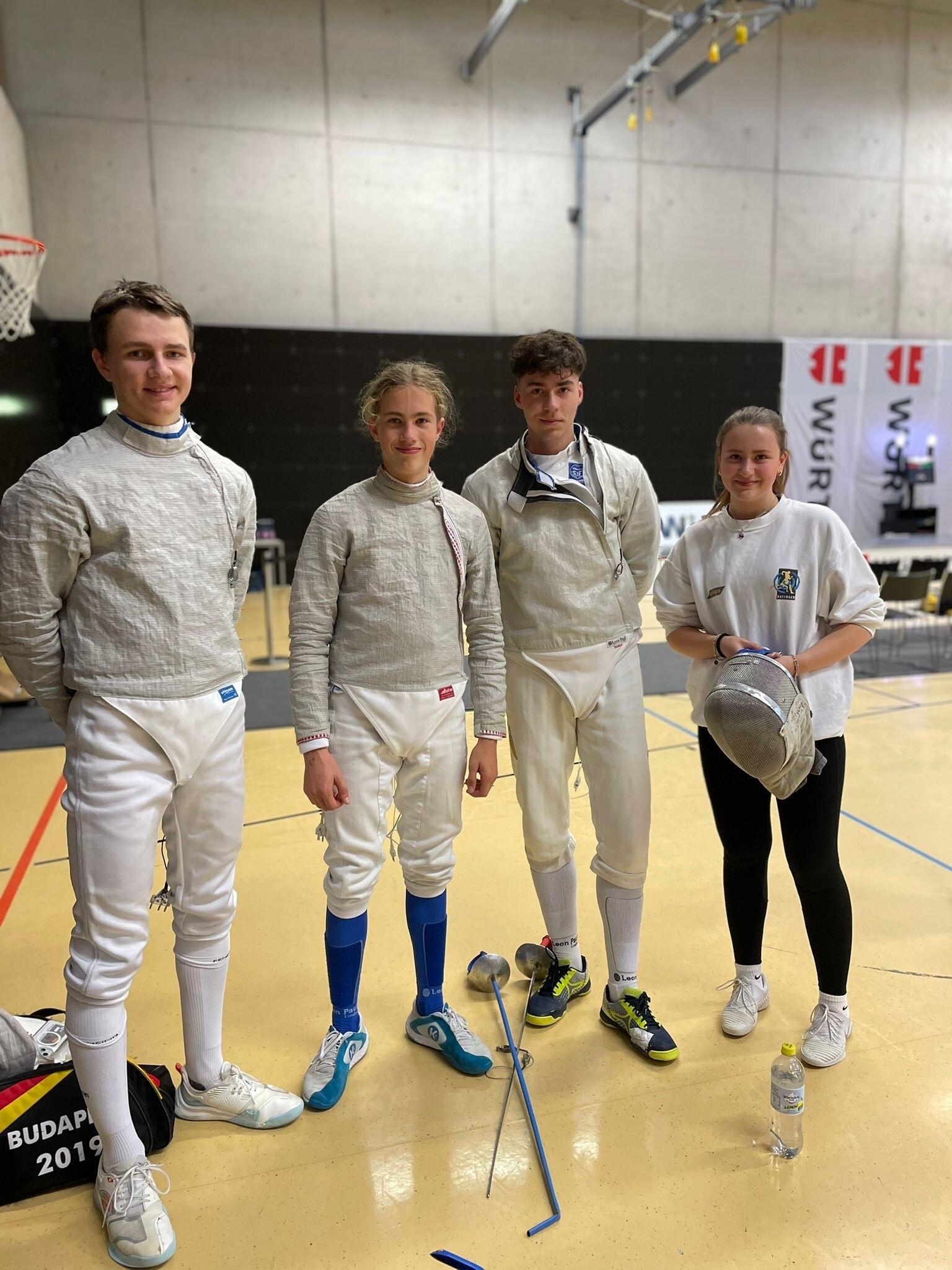 Vier Fechter in Trainingsanzügen stehen in einer Sporthalle, mit Fechtmasken und Ausrüstung um sie herum.