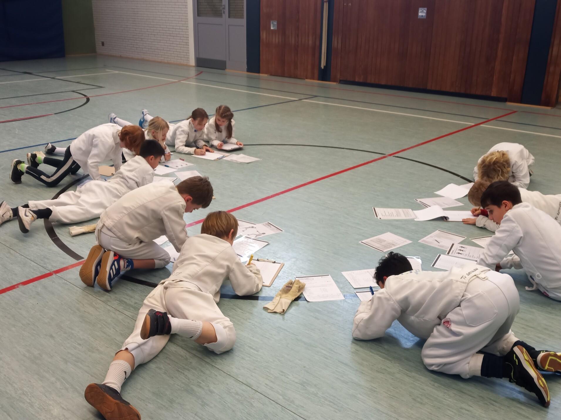 Gruppe von Kindern in Fechtanzügen, die am Boden liegen und konzentriert Arbeitsblätter ausfüllen.