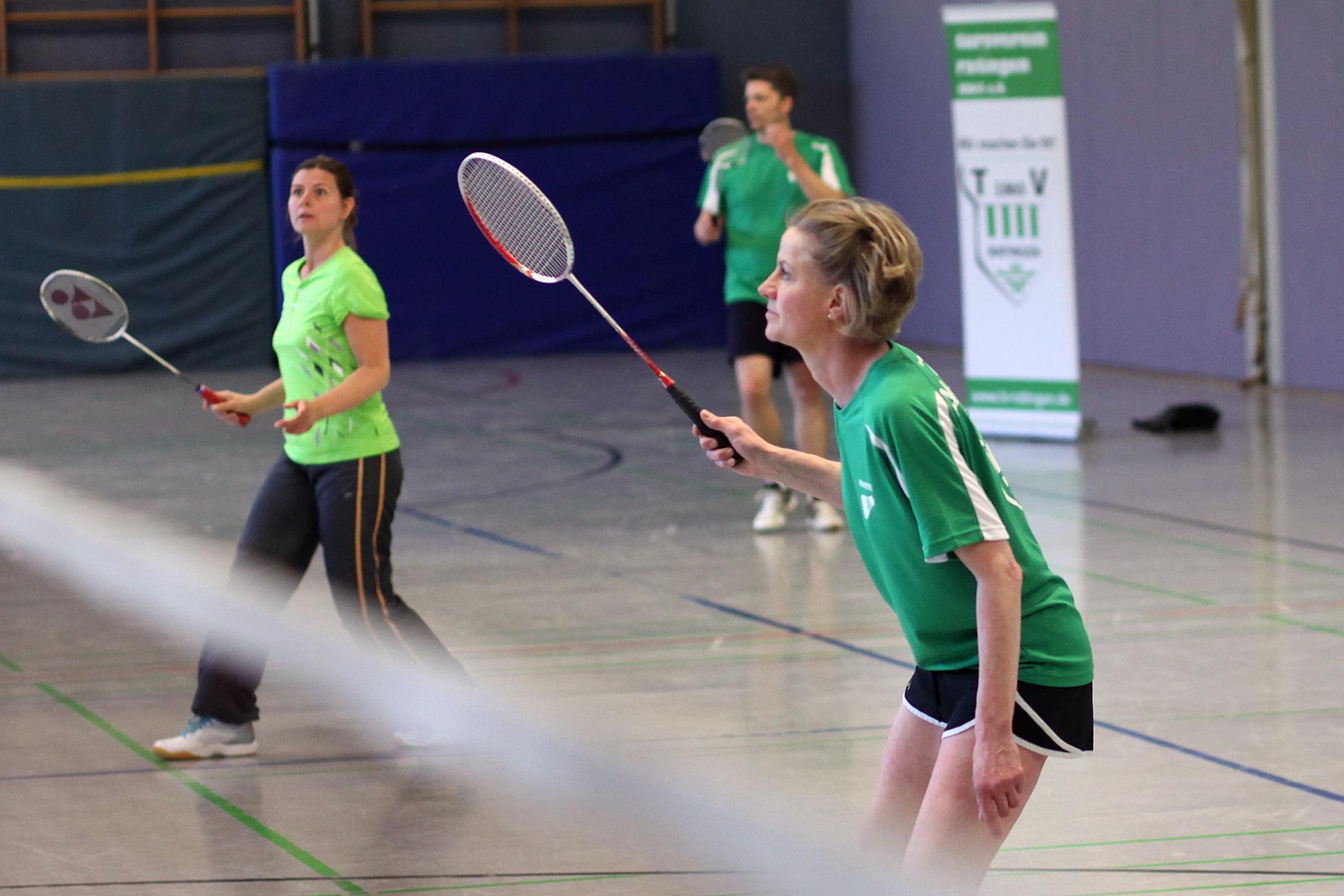 F&uuml;nf Personen in gr&uuml;nen Shirts spielen Badminton in einer Sporthalle. Eines der Netze ist sichtbar.