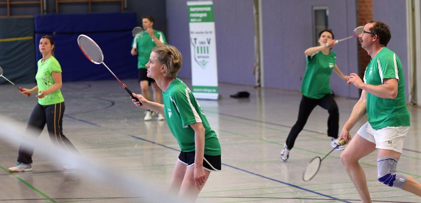 F&uuml;nf Personen in gr&uuml;nen Shirts spielen Badminton in einer Sporthalle. Eines der Netze ist sichtbar.