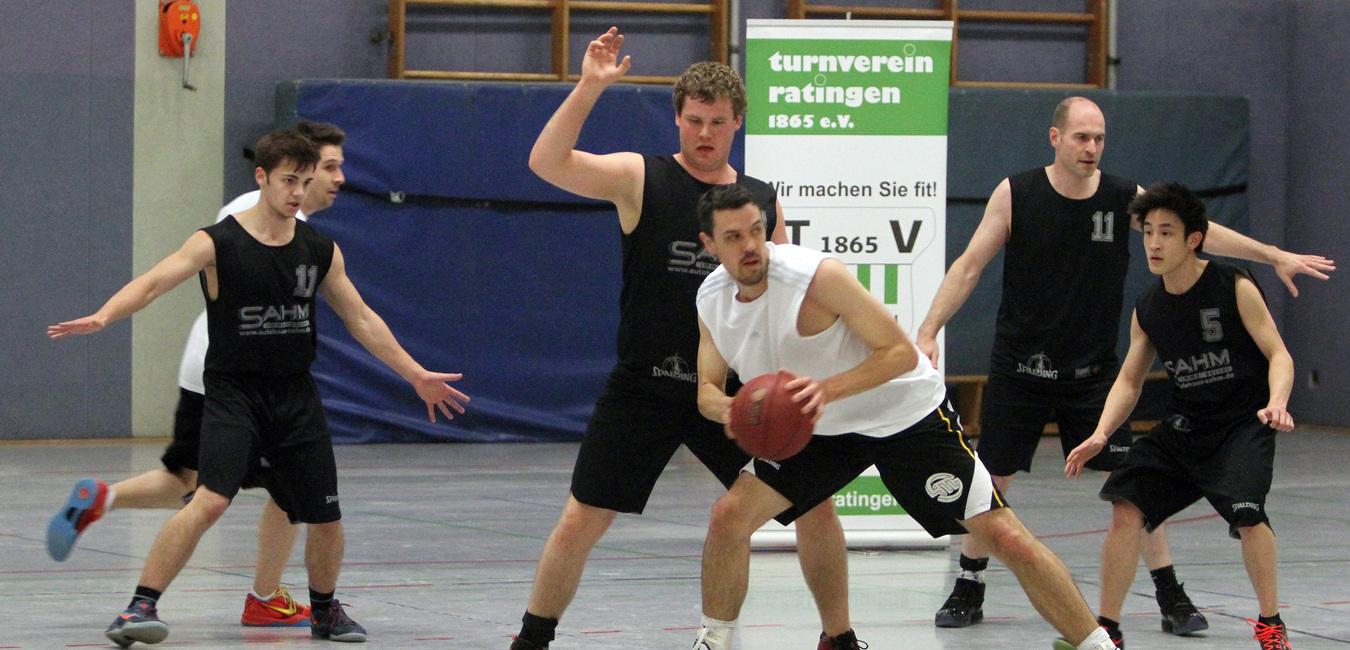Sechs Basketballspieler in Aktion, einer dribbelt den Ball, während die anderen verteidigen. Hintergrund mit Vereinsbanner.