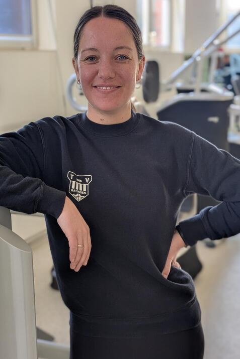 Junge Frau in Sportbekleidung l&auml;chelt und lehnt an einem Fitnessger&auml;t in einem modernen Fitnessstudio.