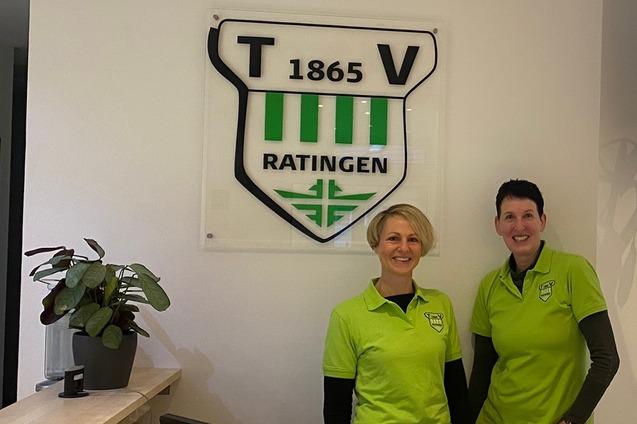 Zwei Mitarbeiterinnen in gr&uuml;nen Poloshirts stehen an einer Empfangstheke vor dem Emblem des T.V. Ratingen.