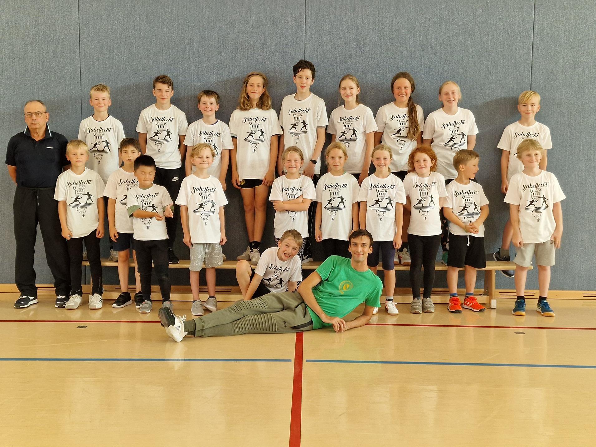 Gruppenfoto von Kindern und Trainern in weißen T-Shirts mit Aufdruck, in einer Sporthalle posierend.
