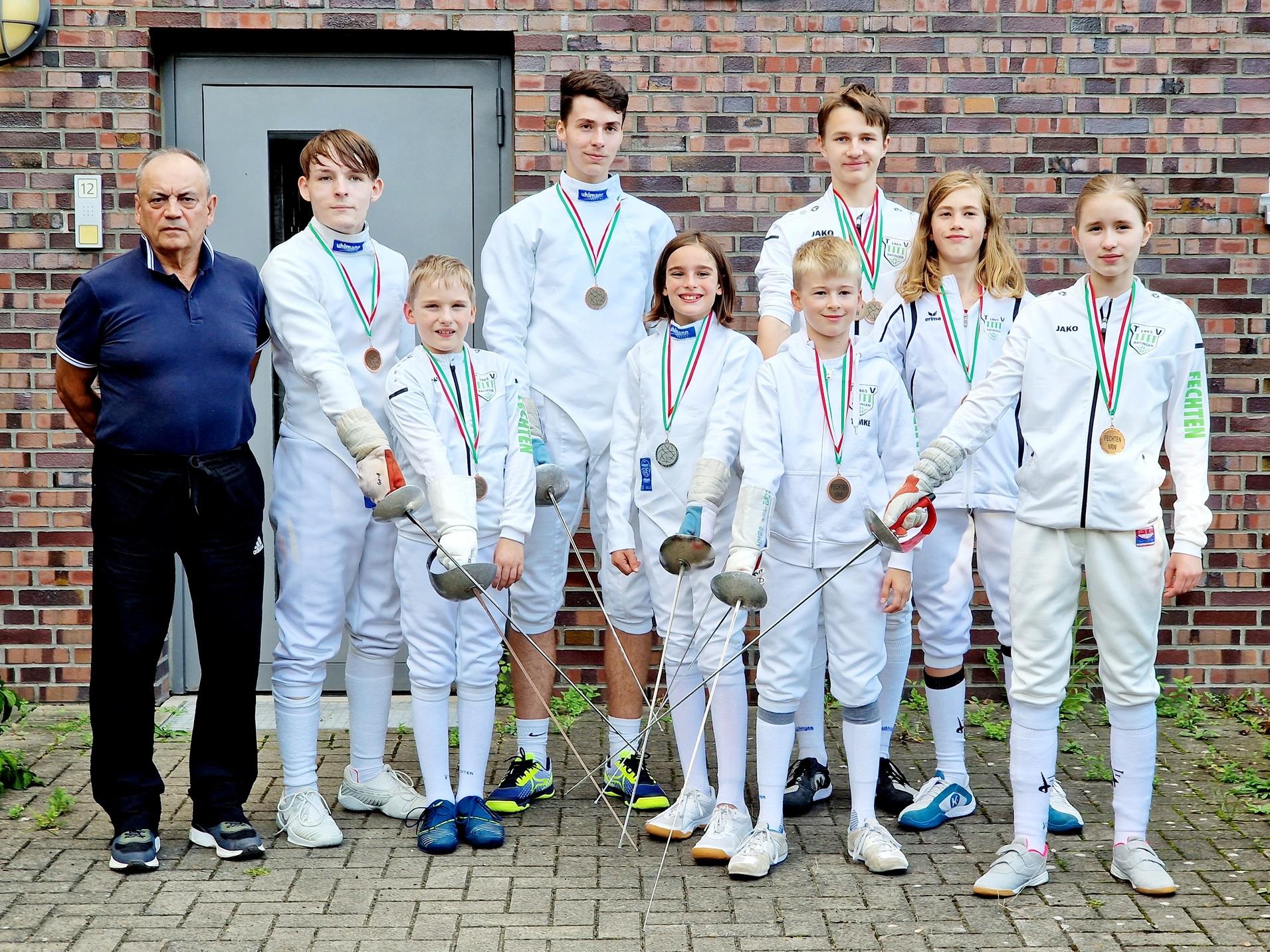 Gruppe von acht Fechtern in weißer Kleidung mit Medaillen, posierend vor einer Backsteinmauer.