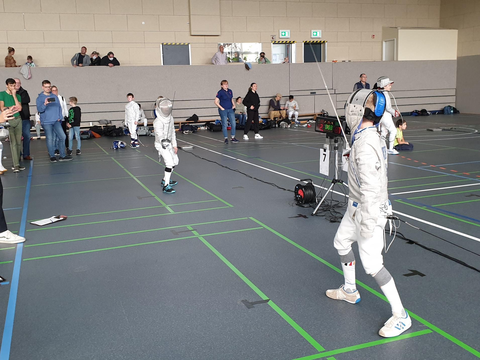 Zwei Fechter in voller Ausrüstung stehen in einer Sporthalle, umgeben von Zuschauern und weiteren Wettkämpfern.