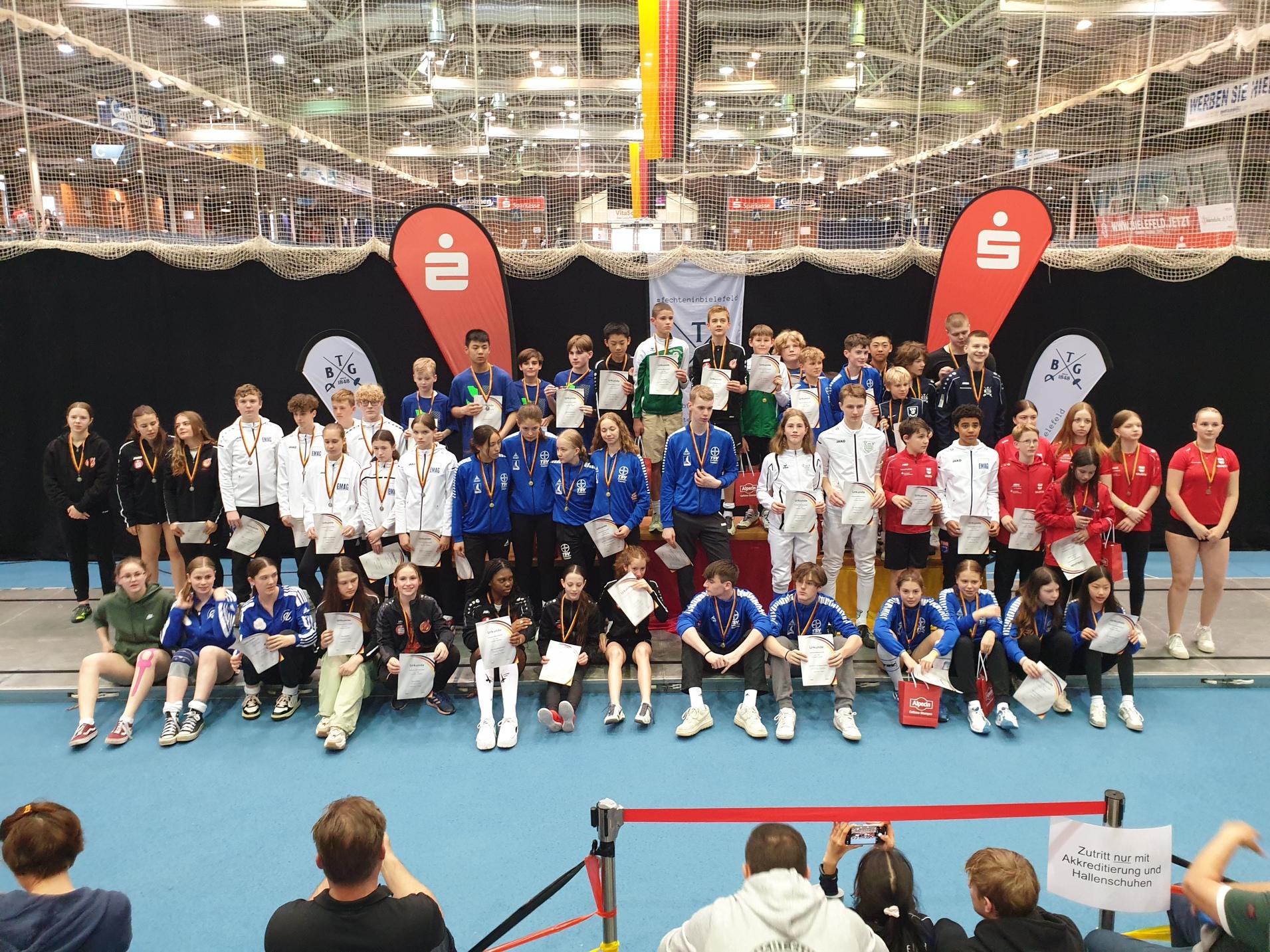 Gruppenfoto von jungen Athleten mit Medaillen und Urkunden auf einem Wettkampf, umgeben von Sponsoren-Logos.