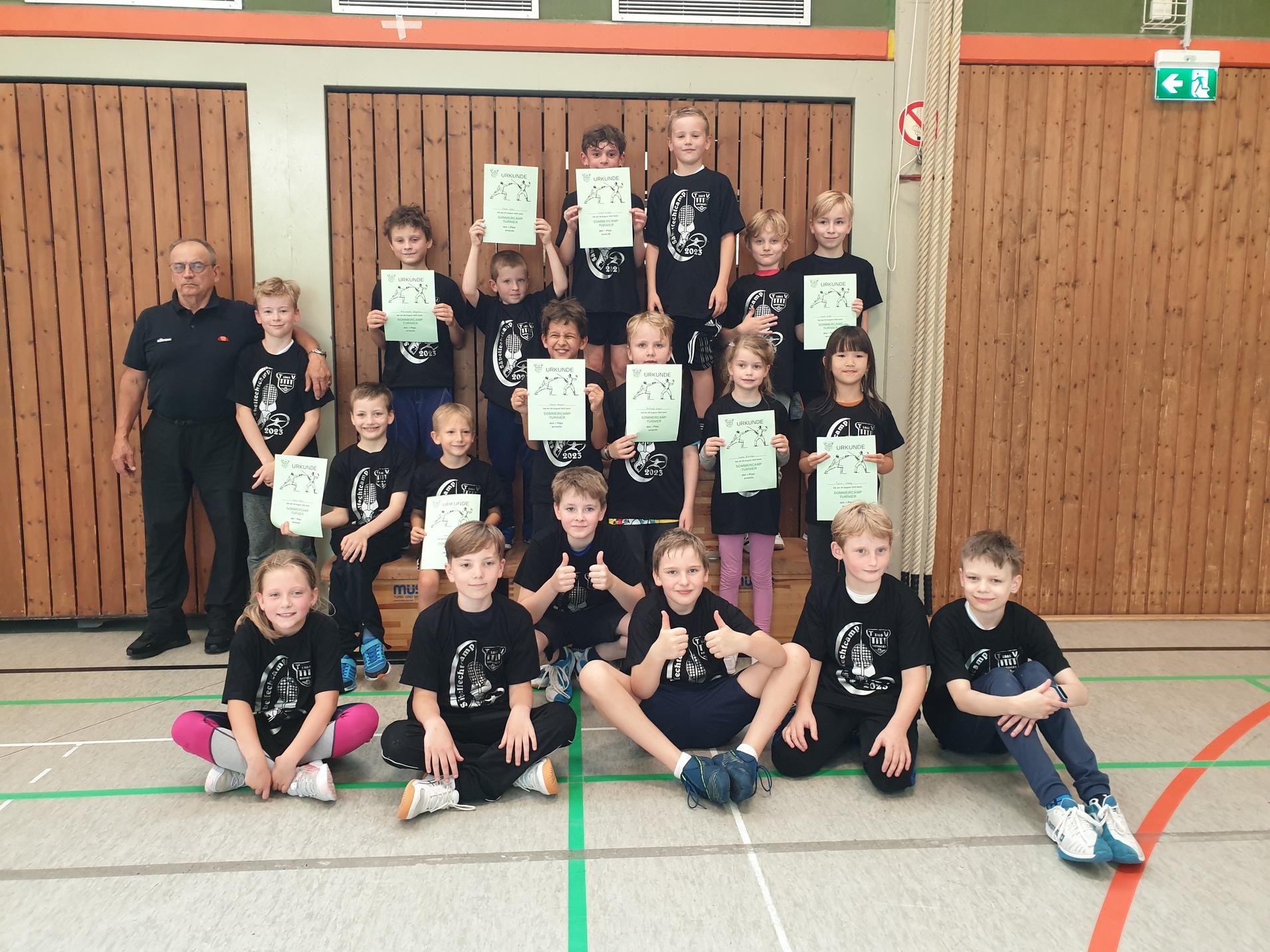Gruppeneinstellung von Kindern in schwarzen T-Shirts, die Urkunden in einer Sporthalle halten, mit einem Erwachsenen auf der linken Seite.