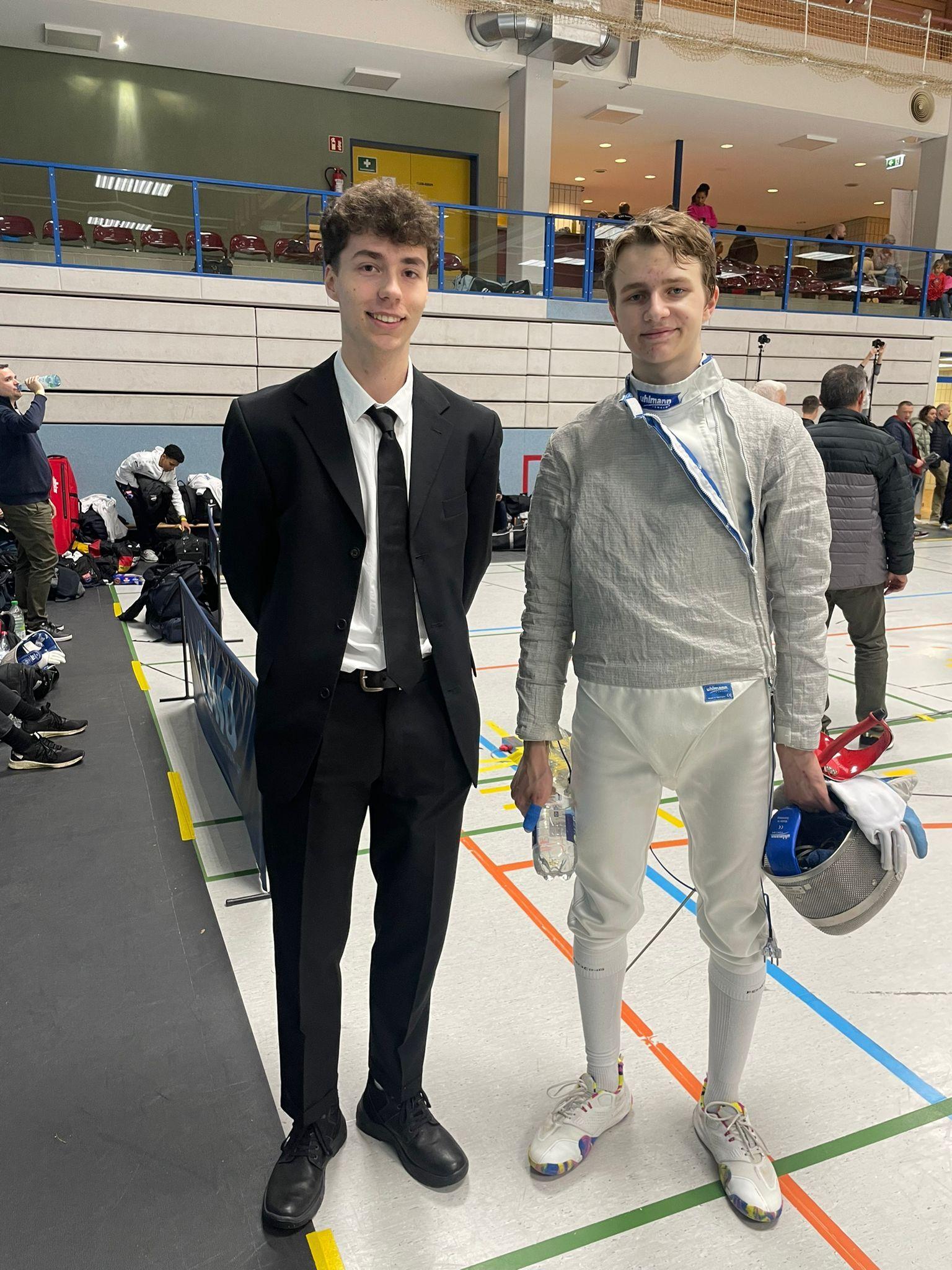 Zwei Jugendliche stehen in einer Sporthalle, einer im Anzug, der andere in Fechtkleidung mit Maske und Handschuh.
