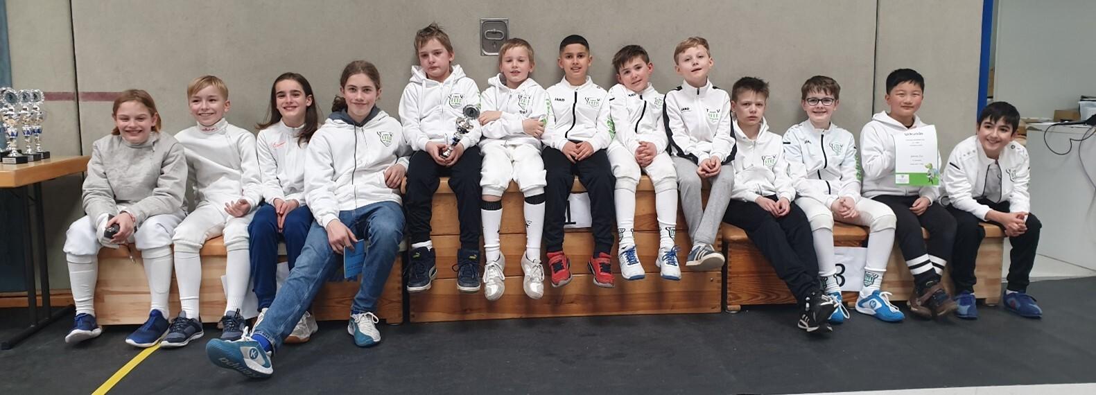 Gruppenschnappschuss von zehn jungen Fechtern in weißen Trainingsjacken, sitzend auf einer Holzbank, mit Pokalen im Hintergrund.