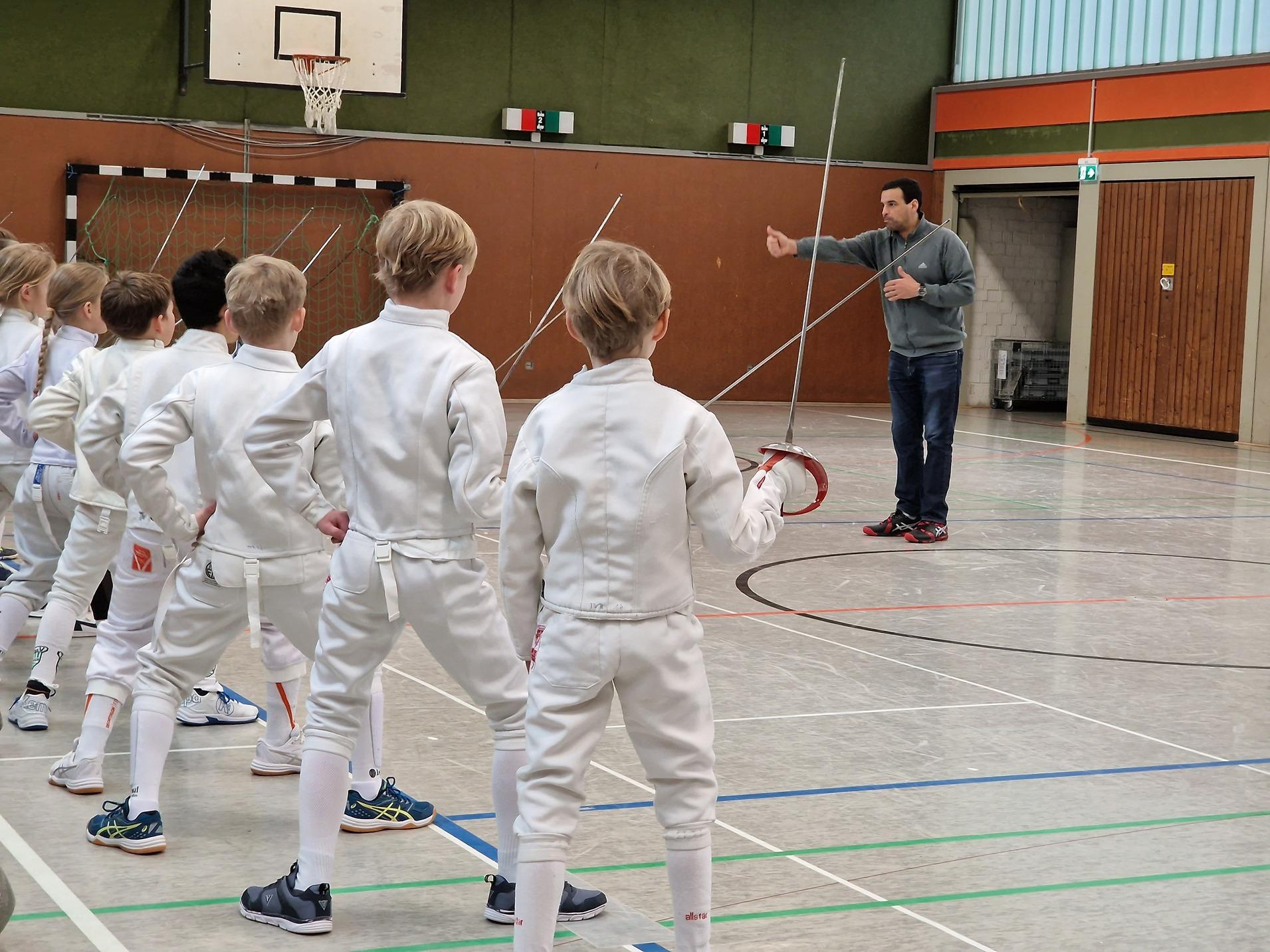 Trainer leitet Gruppe junger Fechter in einer Sporthalle während des Trainings mit Fechtausrüstung.