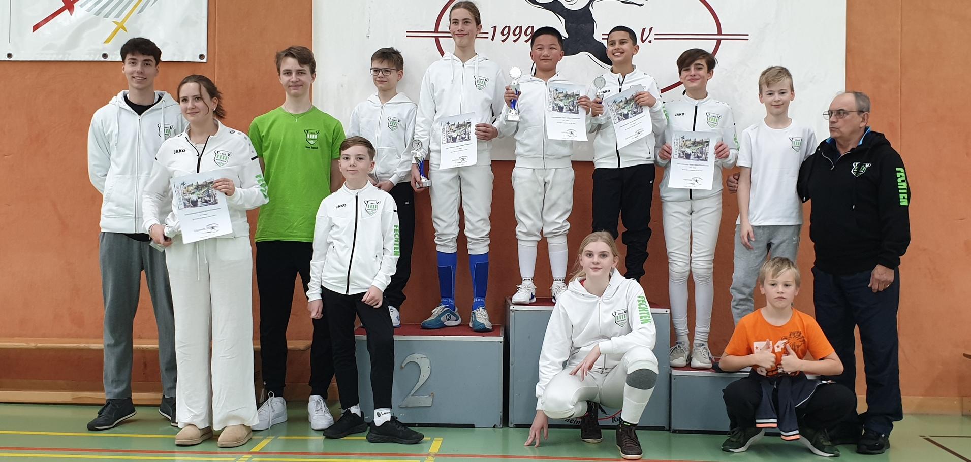 Gruppe von Fechtern in Sportkleidung, posierend mit Urkunden und Pokalen vor einer Vereinswand.