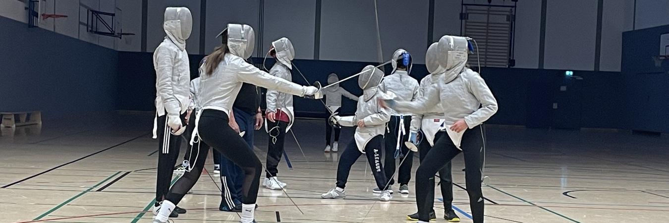 Fünf Fechter in Schutzkleidung üben das Fechten in einer Sporthalle auf einem Holzfußboden.