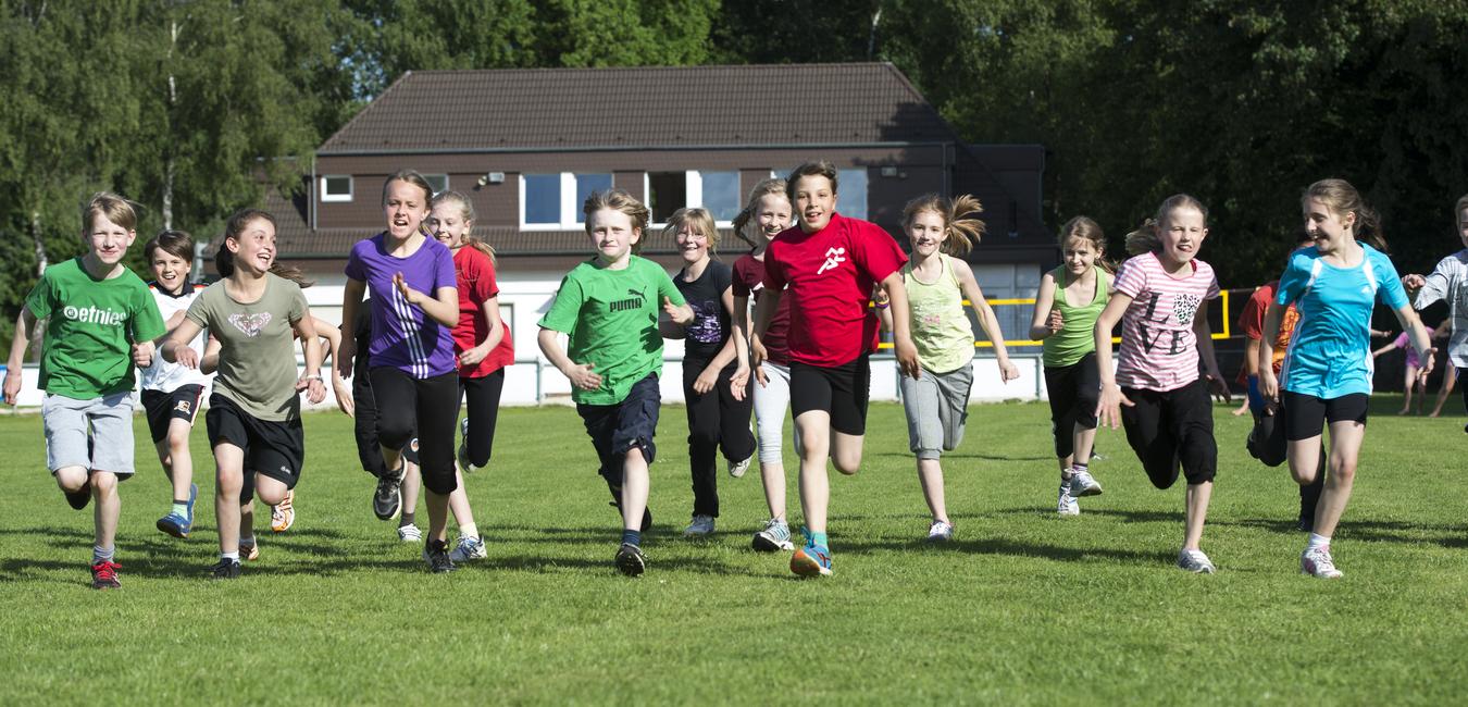 Grupp von Kindern in bunten T-Shirts läuft lachend über ein grünes Sportfeld bei sonnigem Wetter.