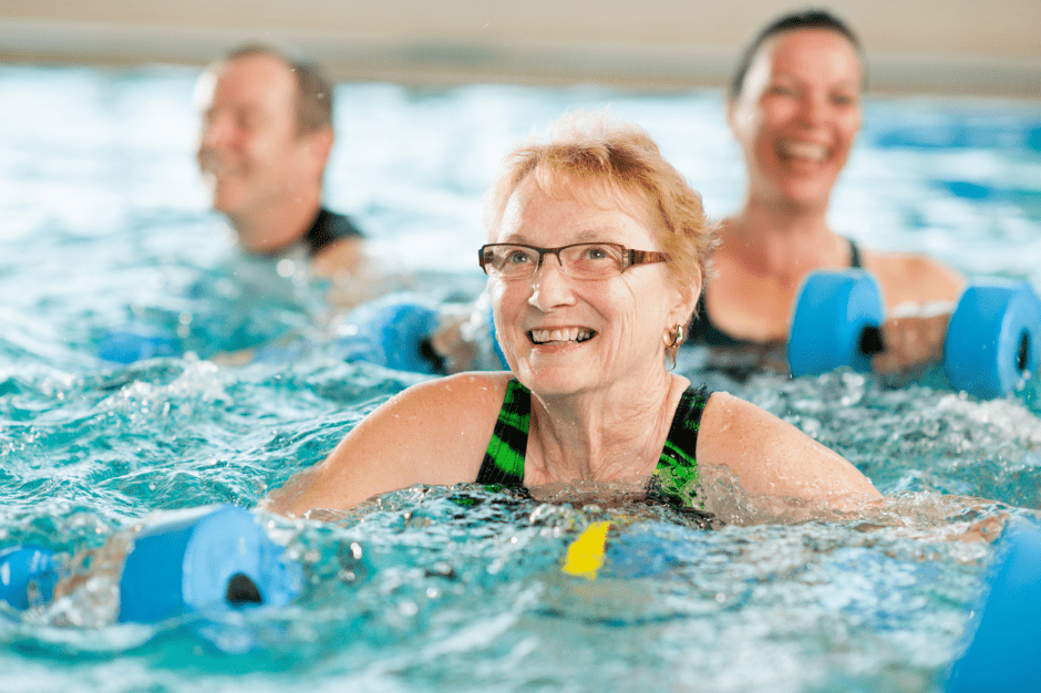 Ältere Frauen und Männer schwimmen mit Wasserbällen in einem hellblauen Pool und lächeln dabei.