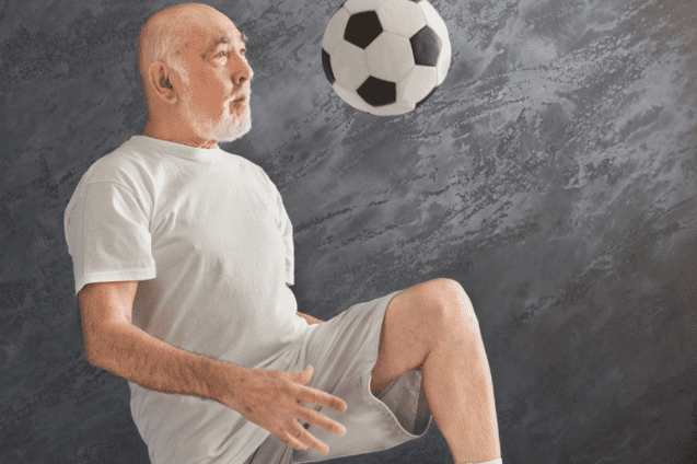 &Auml;lterer Mann in T-Shirt und Shorts jongliert einen Fu&szlig;ball mit dem rechten Bein vor einer grauen Wand.