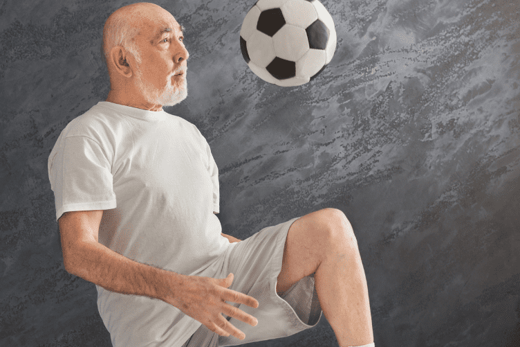 Älterer Mann in T-Shirt und Shorts jongliert einen Fußball mit dem rechten Bein vor einer grauen Wand.