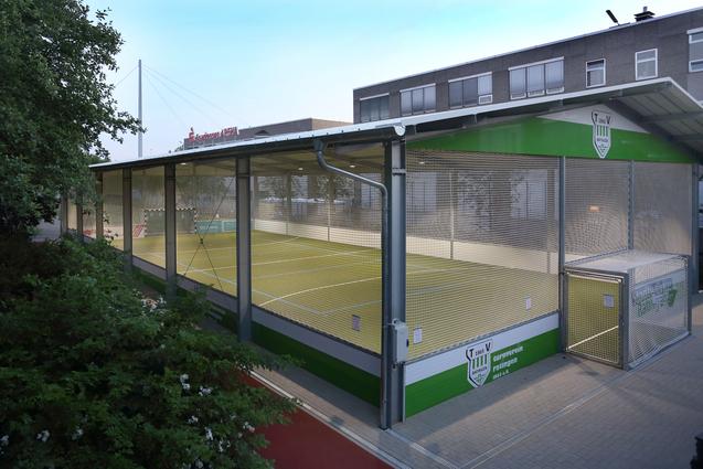 Sicht auf eine &uuml;berdachte Sportanlage mit gr&uuml;ner Spielfl&auml;che und Maschendrahtz&auml;unen, umgeben von B&auml;umen und Geb&auml;uden.