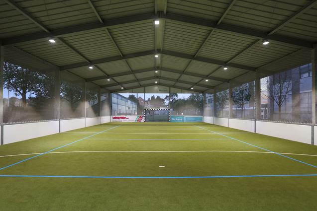 Innenansicht eines Sportfeldes mit Kunstrasen, Beleuchtung und Netzen, umgeben von Fenstern.