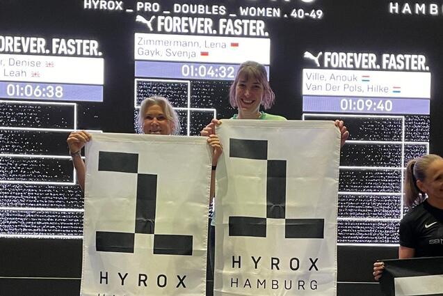 Zwei Siegerinnen halten HYROX-Flaggen auf einem Podium, w&auml;hrend die Ergebnisse im Hintergrund angezeigt werden.