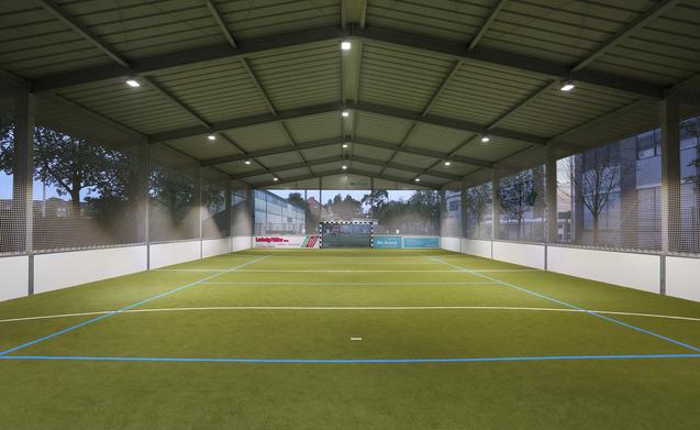 Innenansicht eines Sportfeldes mit Kunstrasen, Beleuchtung und Netzen, umgeben von Fenstern.