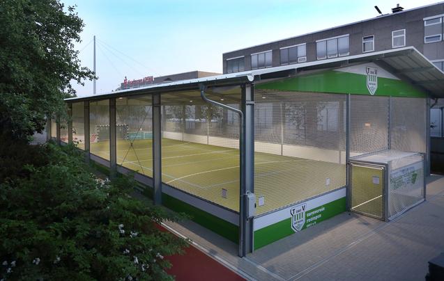 Sicht auf eine überdachte Sportanlage mit grüner Spielfläche und Maschendrahtzäunen, umgeben von Bäumen und Gebäuden.