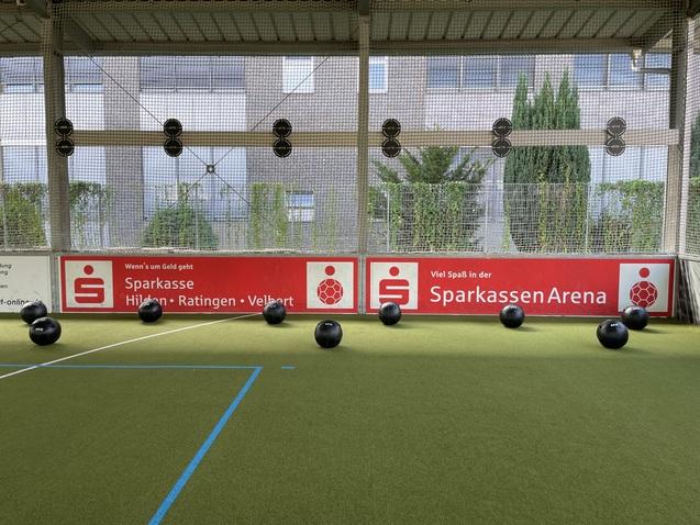 Schwarze Trainingsbälle liegen auf einem grünen Sportboden vor einer Wand mit Werbung für die SparkassenArena.
