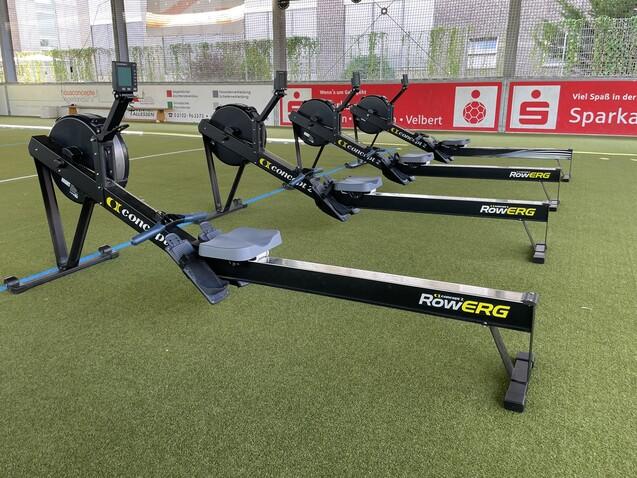 Vier Ruderergometer stehen auf einem Kunstrasenplatz, sichtbar sind Displays und die Ruderzugmechanik.