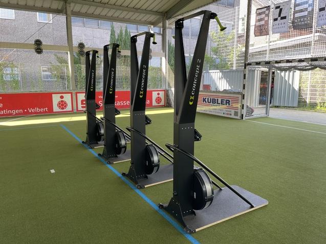 Vier Rudergeräte stehen auf grüner Sportfläche in einer Halle, hinter ihnen sind Werbebanner sichtbar.