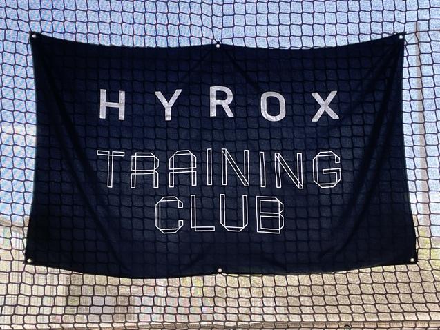 Banner mit der Aufschrift "HYROX TRAINING CLUB" vor einem Netz im Hintergrund.