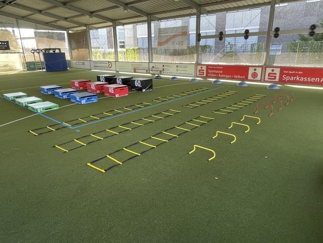 Sportliche Trainingsfläche mit bunten Hürden, Kästen und Leitern auf Kunstrasen in einer Indoor-Arena.
