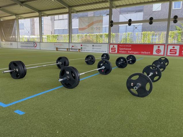 Mehrere Langhanteln mit Gewichten liegen auf einem Sportplatz mit Kunstrasen in einer überdachten Halle.