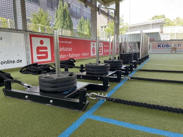 Reihe von Schlitten mit Gewichten und Seilen auf einem Kunstrasenplatz unter einem Netz.