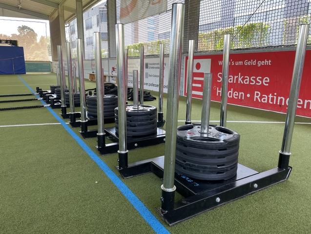 Mehrere Sleds mit Gewichten auf einem Kunstrasenplatz, im Hintergrund eine Wand mit Werbebannern.