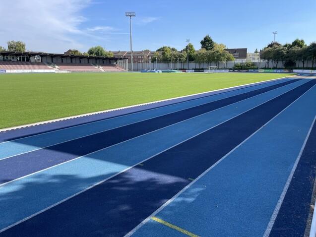 Blick auf einen blauen Laufbahnabschnitt neben einem gr&uuml;nen Fu&szlig;ballfeld in einem Sportstadion.