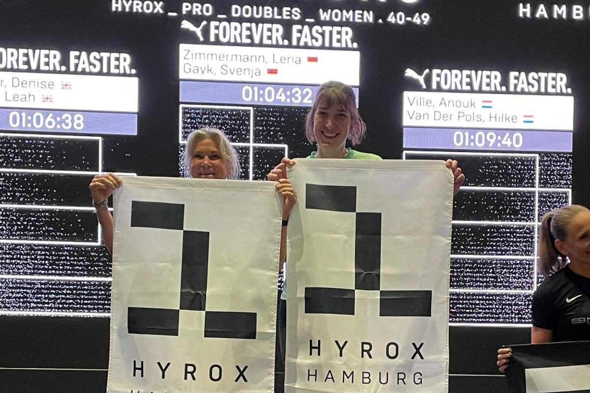 Zwei Siegerinnen halten HYROX-Flaggen auf einem Podium, während die Ergebnisse im Hintergrund angezeigt werden.