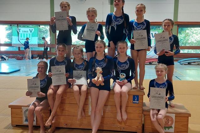 Gruppe junger Turnerinnen in blauen Trikots posiert mit Urkunden auf einem Sprungkasten in einer Turnhalle.