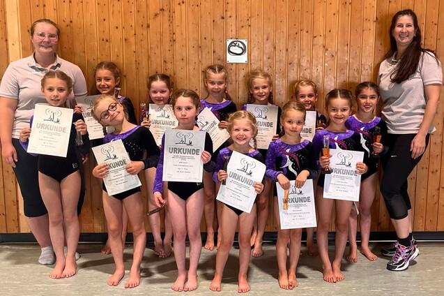 Gruppe von Kindern in Gymnastikanz&uuml;gen h&auml;lt Urkunden, l&auml;chelt vor einer Holzwand mit zwei Trainerinnen.