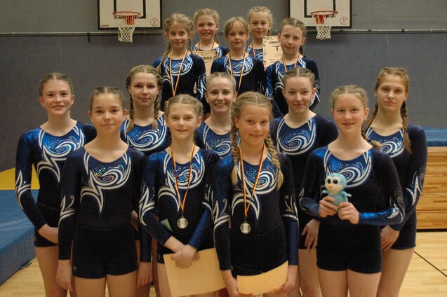 Gruppe von Gymnastinnen in blauen Trikots, mit Medaillen und Urkunden, l&auml;chelnd in einer Sporthalle posierend.